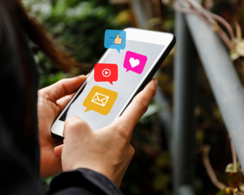A phone being held in a persons hands with social media emoticons.